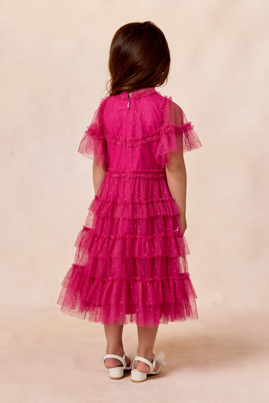 A young girl with long brown hair stands with her back to the camera against a light beige background, wearing the Ivy City Co Mini Whimsical Dress in Fuchsia, which features flutter sleeves and a layered tulle skirt.