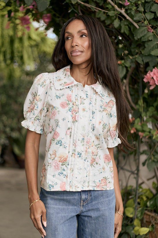 A woman wearing the Ivy City Co Sophia Blouse in Countryside Peony Blossom and blue jeans stands outside near green foliage and pink flowers, looking to the side with a slight smile.