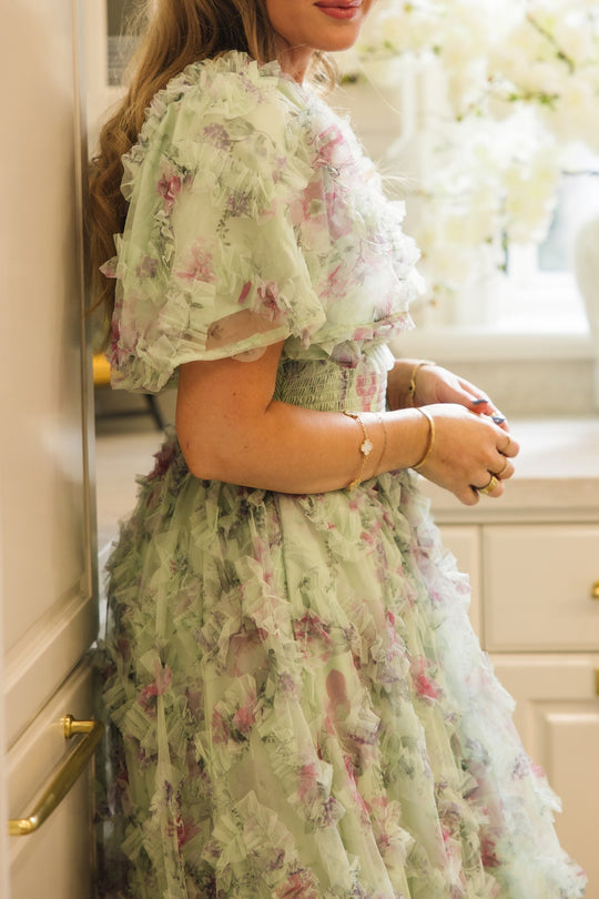 A woman stands by a kitchen counter, hands clasped, wearing the Florentina Dress in Pistachio Aquarelle Fleur by Ivy City Co. The elegant nursing-friendly dress features pistachio floral chiffon with ruffled, sheer fabric. She accessorizes with gold bangles and a ring as light pours through the window, illuminating white flowers in the background.
