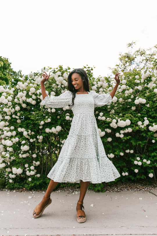 A woman wearing the Madeline Dress in Blue by Ivy City Co, featuring a light blue floral print and smocked bodice, smiles and poses with her arms raised in front of a lush bush covered in white flowers on a sunny day.