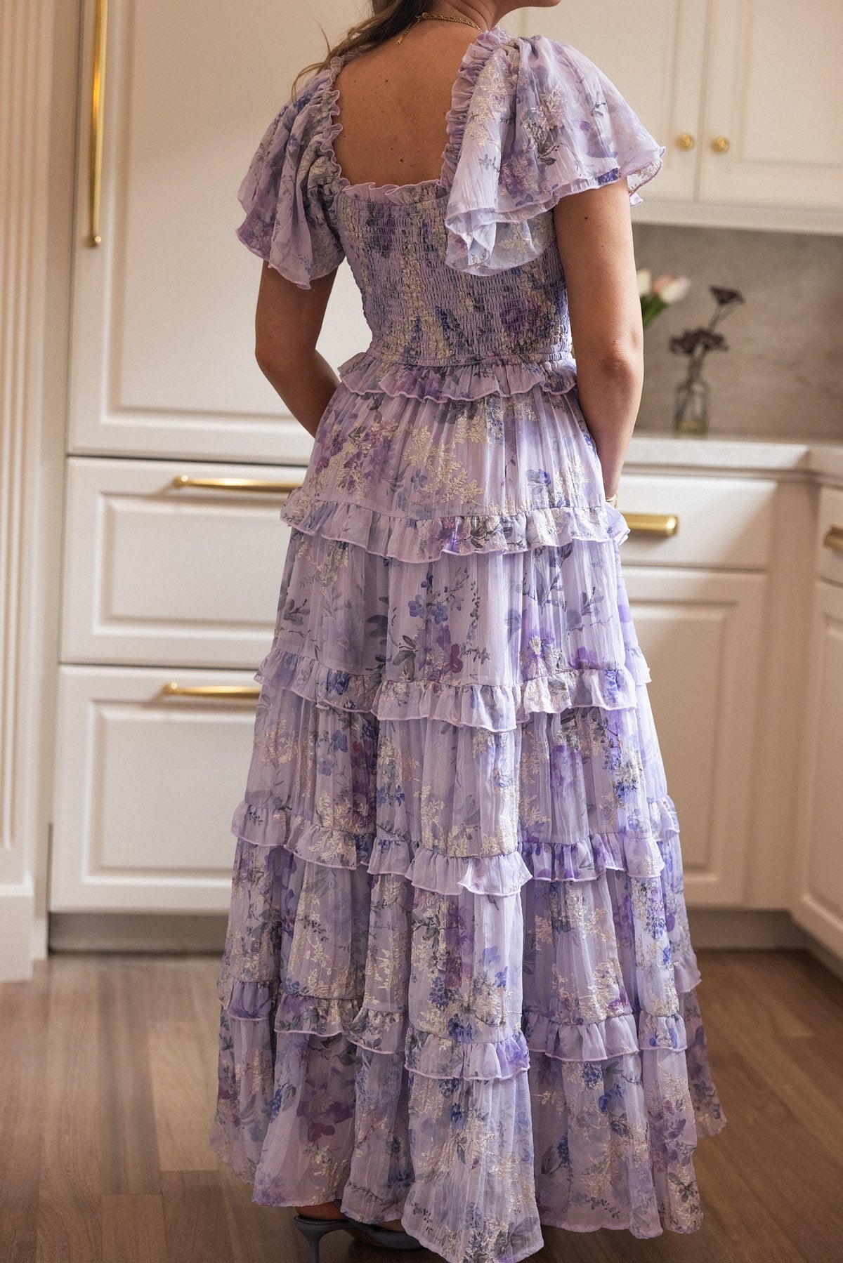 A person stands in a kitchen facing away from the camera, wearing the Winnie Dress in Lavender Aquarelle Fleur by Ivy City Co—a graceful, maternity-friendly chiffon dress featuring floral ruffles, short puff sleeves, and tiered layers, perfect for capturing the soft glow of spring.