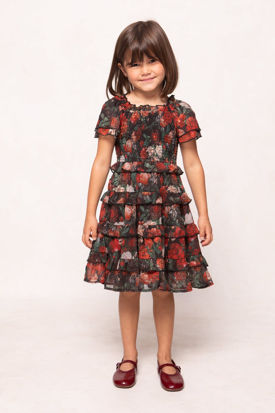 A young girl with straight brown hair stands and smiles against a plain light background, wearing the Ivy City Co Mini Winnie Dress in Midnight Roses—a floral girls’ maxi dress with ruffled layers and short sleeves. She completes her look with maroon shoes.
