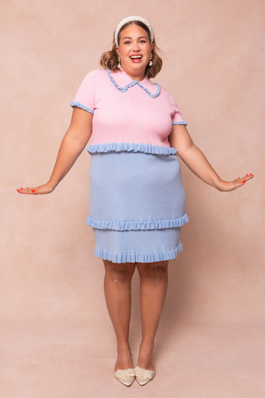 A smiling woman poses with arms outstretched, wearing Ivy City Cos Courtney Knit Dress in pink with a blue collar and ruffled skirt. She accessorizes with a headband, hoop earrings, pointed shoes, and stands against a beige background.