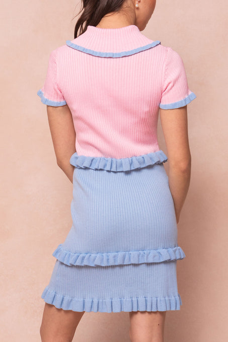 A woman stands facing away, wearing the Ivy City Co Courtney Knit Dress. This maternity-friendly dress features a ribbed pink short-sleeve top, light blue skirt, and blue ruffle trim against a beige background.