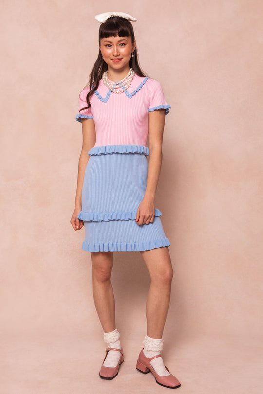 A woman smiles against a beige background wearing the Ivy City Co Courtney Knit Dress in pastel pink and blue with ruffled trim, white socks, pink shoes, layered pearl necklaces, and a large white hair bow. This look is maternity friendly.
