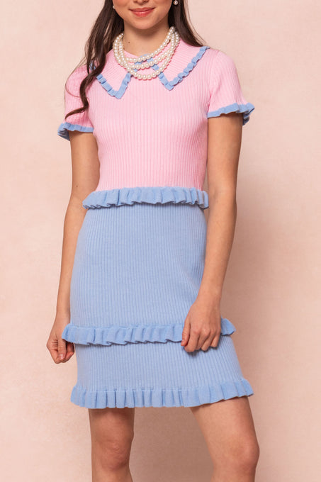 A woman models the Ivy City Co Courtney Knit Dress, a maternity-friendly, ribbed pink short-sleeve dress with blue ruffled trim and matching skirt, styled with layered pearl necklaces against a light pink backdrop.