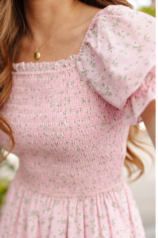 A woman wears the Ivy City Co Valencia Dress in Pink Ditsy Floral, a maternity-friendly style featuring puff sleeves and a smocked bodice, paired with a gold pendant necklace. Her face is out of frame.