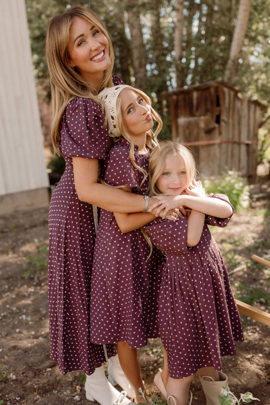 Three people—a woman and two girls—stand outdoors hugging and smiling, all wearing the Ivy City Co Teen Blakely Dress in Aubergine Ditsy Dot. Trees and a wooden shed can be seen in the background.