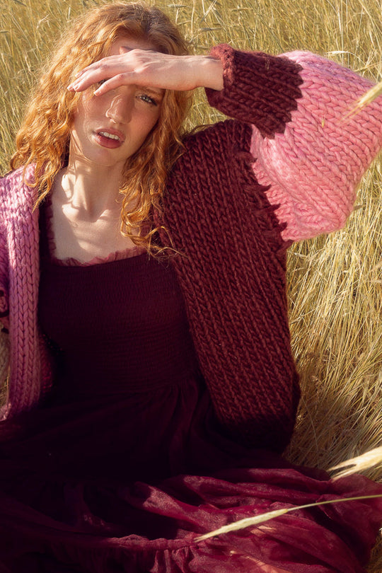 A woman with red hair sits in tall grass, wearing a burgundy dress and the Marissa Sweater in Pink & Red by Ivy City Co, shielding her eyes from the sun with her hand.