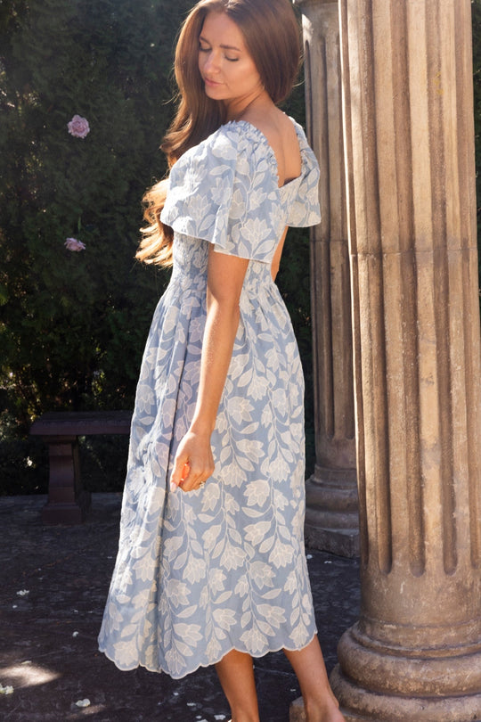 Wearing the Ophelia Dress in Mosaic Embroidery by Ivy City Co, a woman stands beside a stone column outdoors, sunlight illuminating her long brown hair and the elegant embroidered details of her dress.