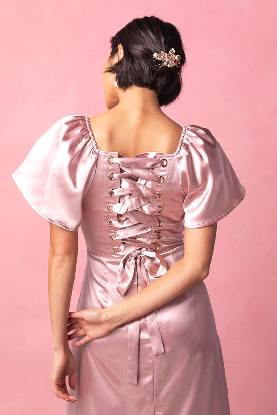 A woman with short dark hair stands with her back to the camera against a pink background, wearing the Ivy City Co Maddie Dress in Blush Satin, featuring puff sleeves and a lace-up back. She accessorizes her look with a decorative hairpin.