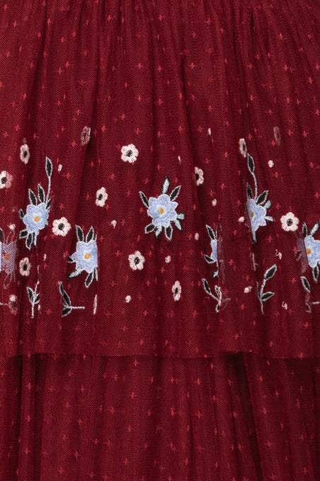 Close-up of the Ivy City Co Mabel Dress in Burgundy, a maxi dress with layered tulle skirt, detailed with blue and white floral embroidery, green leaves, and delicate white dots in a decorative pattern.