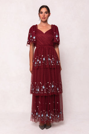 A woman stands against a light background wearing the Ivy City Co Mabel Dress in Burgundy, featuring blue floral embroidery, a layered tulle skirt, and short flutter sleeves. Her hair is pulled back, and she finishes the look with pointed shoes.