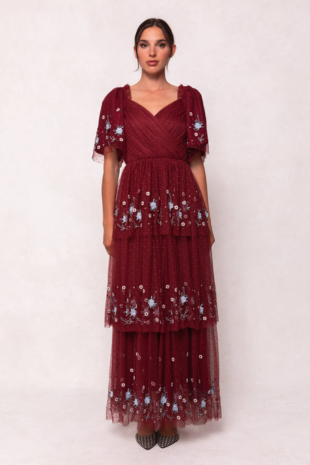 A woman stands against a light background wearing the Ivy City Co Mabel Dress in Burgundy, featuring blue floral embroidery, a layered tulle skirt, and short flutter sleeves. Her hair is pulled back, and she finishes the look with pointed shoes.
