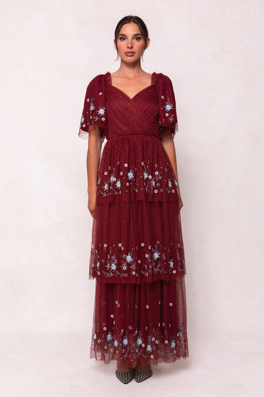 A woman stands against a light background wearing the Ivy City Co Mabel Dress in Burgundy, featuring blue floral embroidery, a layered tulle skirt, and short flutter sleeves. Her hair is pulled back, and she finishes the look with pointed shoes.
