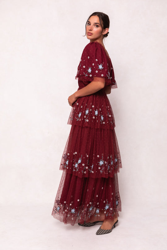 A woman stands sideways in the Ivy City Co Mabel Dress in Burgundy, featuring a tiered maroon tulle skirt and blue and white floral embroidery. She wears her hair back and pairs the dress with black and white checkered shoes against a plain background.