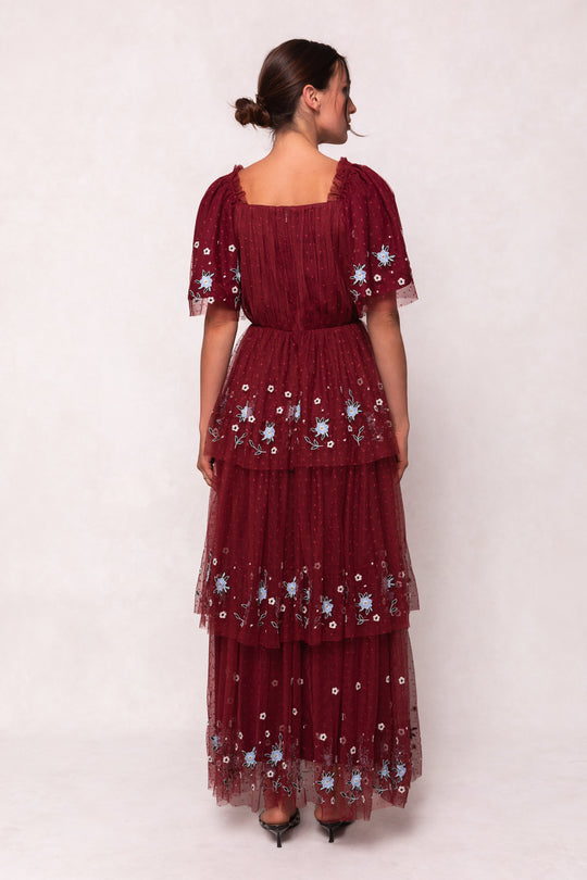 A woman, hair in a low bun, models the Ivy City Co Mabel Dress in Burgundy—a short-sleeved maxi with blue and white floral embroidery and a layered tulle skirt—paired with black heels against a light background.