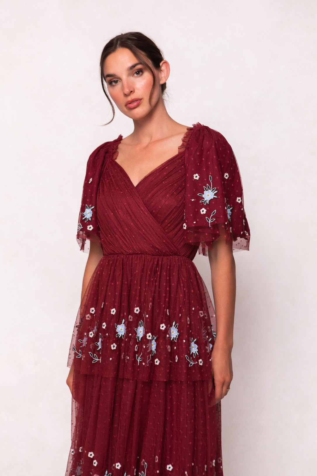 A woman stands against a plain light background wearing the Ivy City Co Mabel Dress in Burgundy, featuring blue and white floral embroidery, sheer puffed short sleeves, and a wrap-style v-neckline.