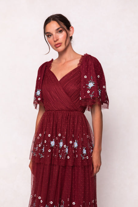 A woman stands against a plain light background wearing the Ivy City Co Mabel Dress in Burgundy, featuring blue and white floral embroidery, sheer puffed short sleeves, and a wrap-style v-neckline.