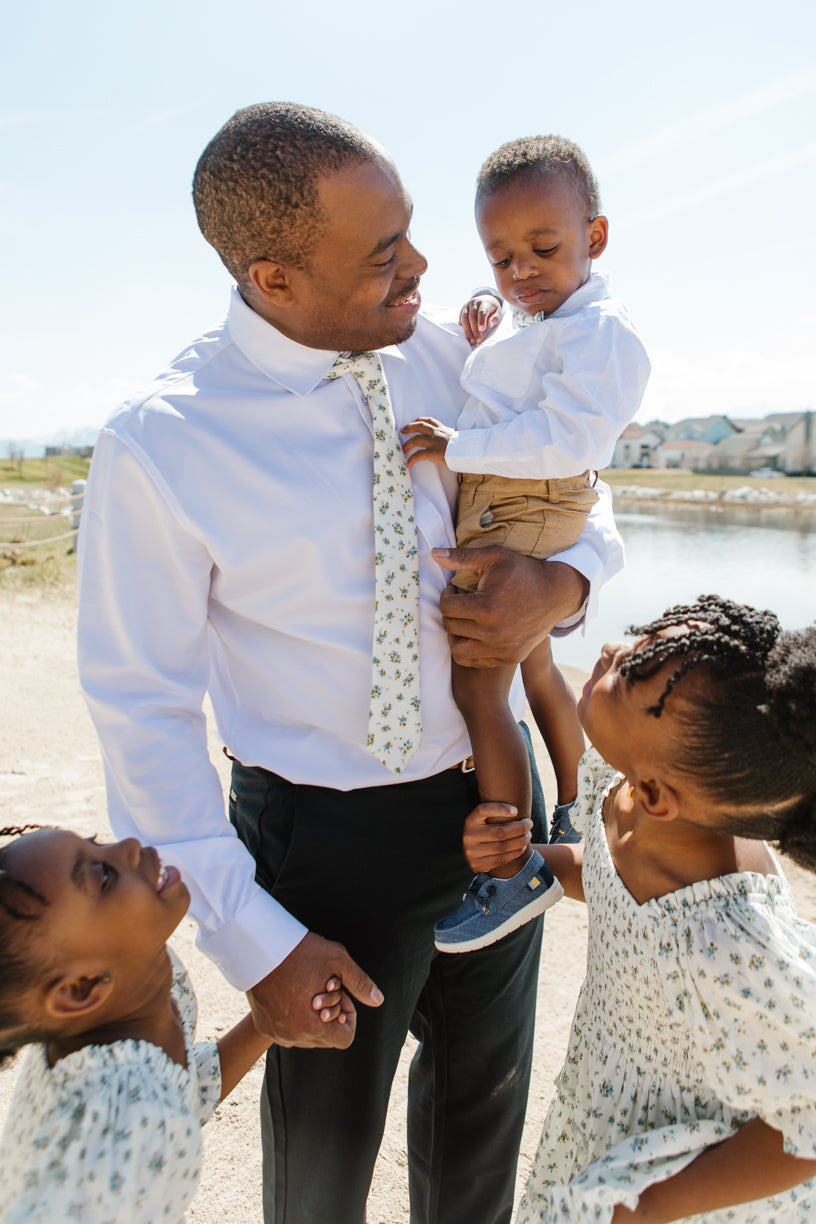A man in a white shirt and an Ivy City Co Madeline Blue Floral tie smiles, holding a boy in a bow tie. Two girls in matching Madeline Blue Floral dresses by Ivy City Co look up at them outdoors near water and houses on a sunny day.