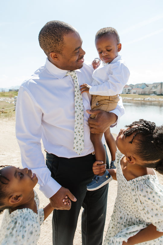 A man in a white shirt and an Ivy City Co Madeline Blue Floral tie smiles, holding a boy in a bow tie. Two girls in matching Madeline Blue Floral dresses by Ivy City Co look up at them outdoors near water and houses on a sunny day.