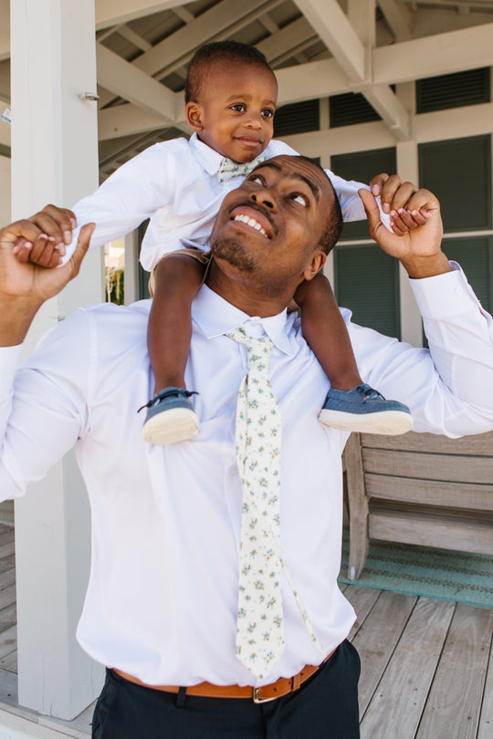A man in a white shirt and Ivy City Co’s Ties in Madeline Blue Floral smiles at a child on his shoulders. The child, wearing a white shirt and blue shoes, holds the man’s hands as they stand under an outdoor wooden structure.