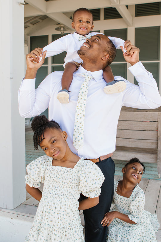 A man in a white shirt and Ivy City Co Ties in Madeline Blue Floral smiles while carrying a boy with a bow tie on his shoulders. In front, two girls in matching floral dresses pose—they all beam happily outside a building.
