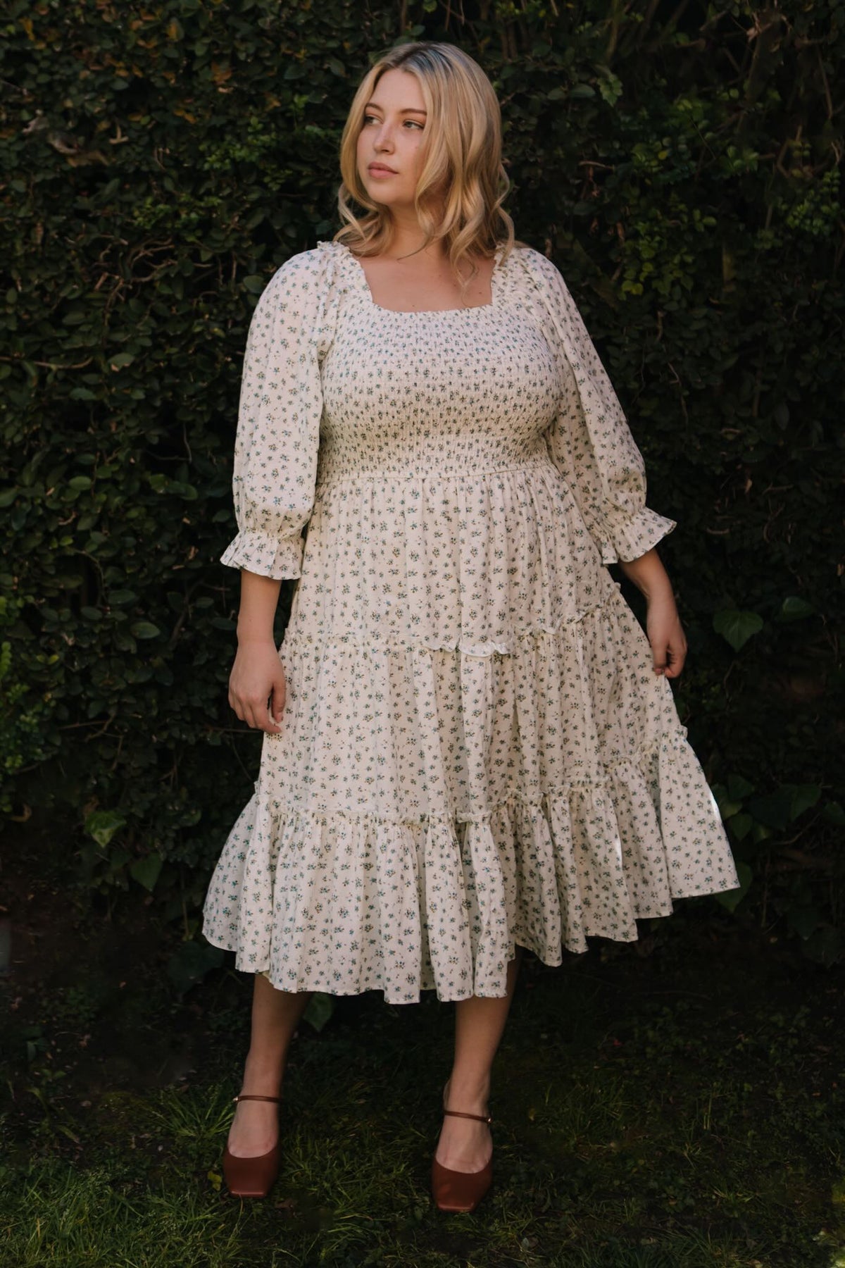 A blonde woman stands outdoors before green bushes, wearing the Ivy City Co Madeline Dress in Blue—a floral, smocked midi that's maternity and nursing friendly—paired with brown platform shoes. She gazes calmly to the side.