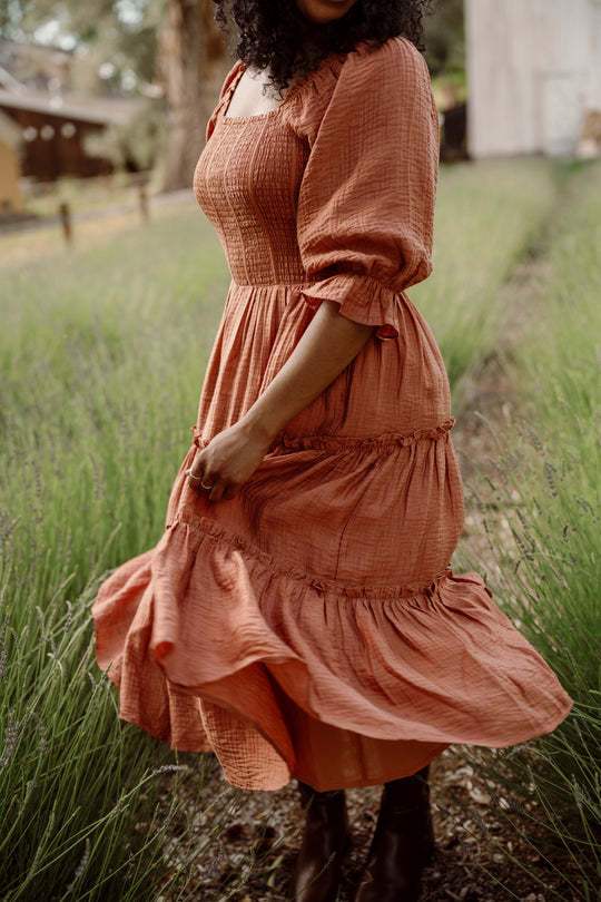 A person twirls in a field of green grass, wearing the Ivy City Co Madeline Dress in Apple Cider—a flowing, rust-colored style with a smocked bodice—while rustic buildings blur in the background. This dress is also maternity friendly, providing both comfort and elegance.