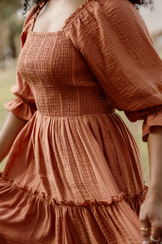 An individual wearing the Ivy City Co Madeline Dress in Apple Cider—a rust-colored, textured dress featuring puffed sleeves and a smocked bodice—stands outdoors, holding the skirt. The focus is on the dress and torso, with the face not shown. This style is also maternity friendly.