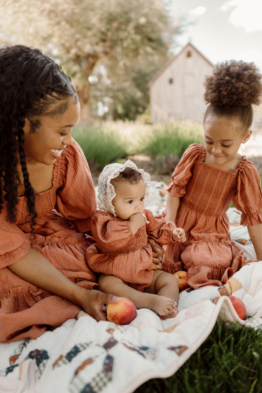 A woman and two young girls, all dressed in matching Ivy City Co Madeline Dresses in Apple Cider featuring charming smocked bodices, sit on a blanket outdoors with peaches. The baby wears a lace bonnet. Trees and a rustic barn appear in the background, enhancing the maternity-friendly setting.