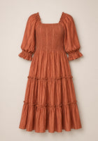 The Madeline Dress in Apple Cider by Ivy City Co features a rust-colored, knee-length silhouette with puffed three-quarter sleeves, a maternity-friendly smocked bodice, and a tiered, ruffled skirt, shown against a neutral background.