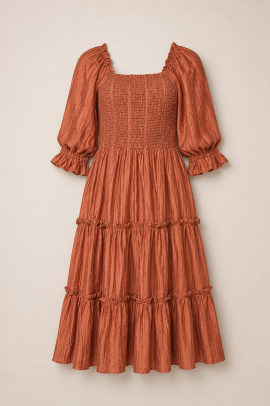 The Madeline Dress in Apple Cider by Ivy City Co features a rust-colored, knee-length silhouette with puffed three-quarter sleeves, a maternity-friendly smocked bodice, and a tiered, ruffled skirt, shown against a neutral background.