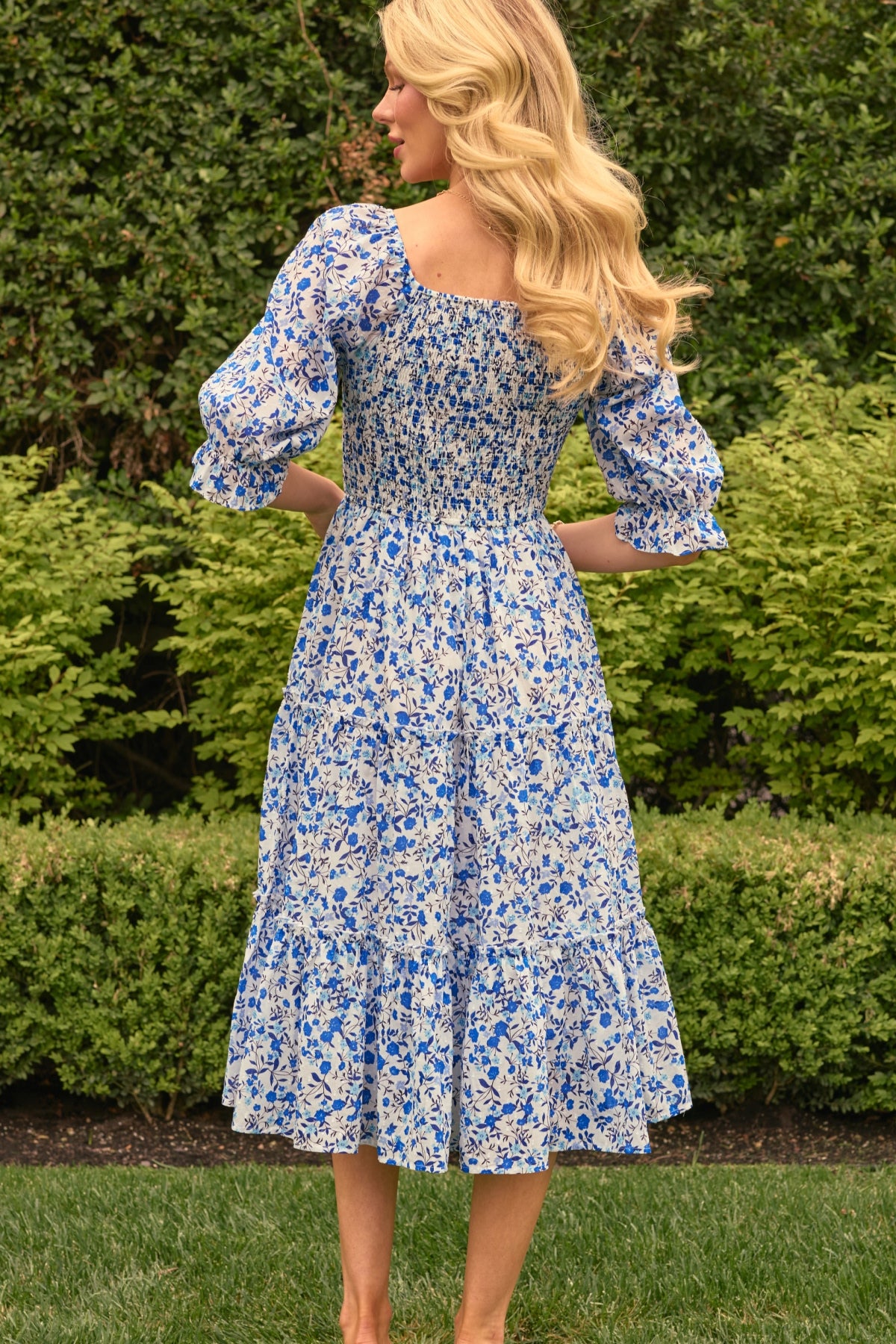 A woman with long blonde hair stands on grass, facing away, wearing the Ivy City Co Madeline Dress in Ditsy Cobalt Floral—a blue and white cotton midi that’s maternity and nursing friendly—amid green bushes and trees.
