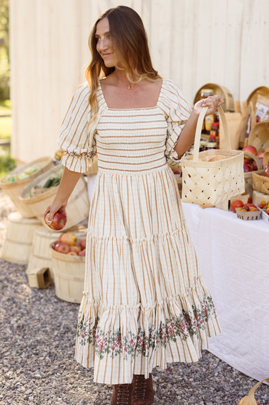 A woman in Ivy City Co’s Madeline Dress in Embroidered Cider Grid, featuring a smocked bodice and floral trim, holds apples and a basket at an outdoor market. Behind her, baskets brimming with apples rest on a white-covered table, highlighting the dress’s charming, maternity-friendly design.