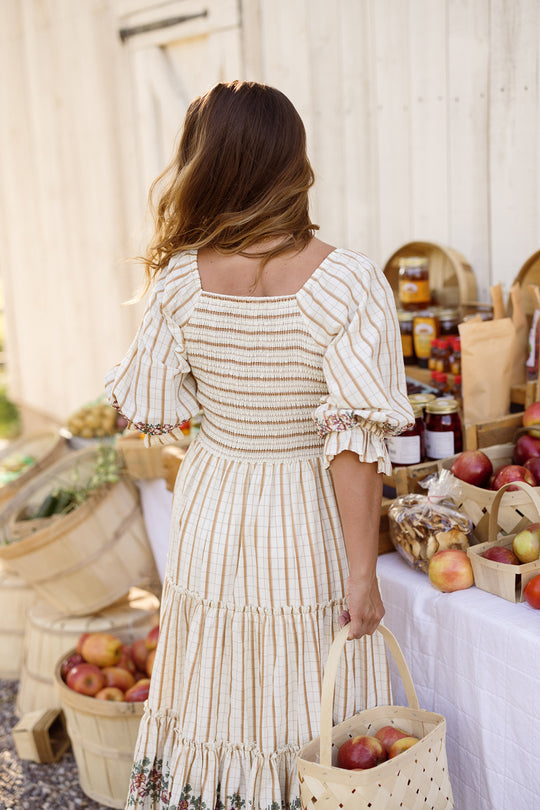 Dressed in Ivy City Co’s Madeline Dress in Embroidered Cider Grid, a woman stands near a white wooden wall at an outdoor market table filled with apples, jars, and produce baskets. She holds a basket and faces away from the camera, creating a charming and maternity-friendly look.