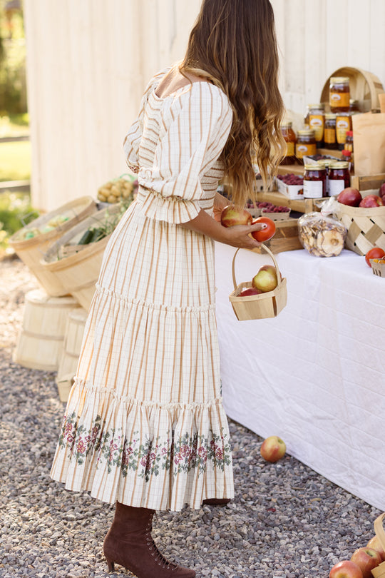 Dressed in the Madeline Dress in Embroidered Cider Grid by Ivy City Co—a relaxed-fit, maternity-friendly style with a smocked bodice—a woman in brown boots picks apples at an outdoor market stand filled with baskets of produce and jars of preserves displayed on a white table. This comfortable and stylish dress is perfect for a day spent gathering fresh finds.
