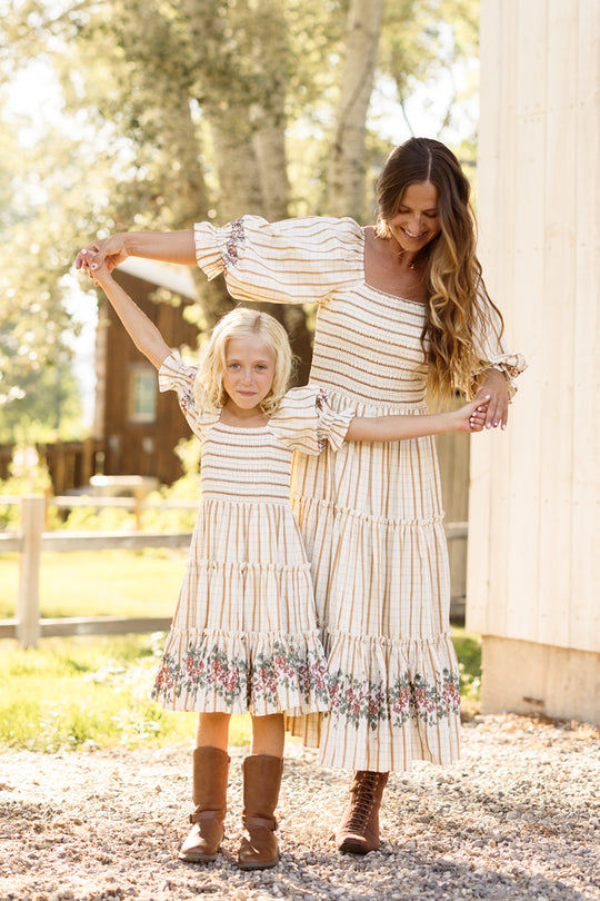 A woman and a young girl hold hands and smile outdoors on a sunny day, both wearing the Ivy City Co Madeline Dress in Embroidered Cider Grid—a matching style with smocked bodices that’s even maternity friendly for every stage of motherhood.