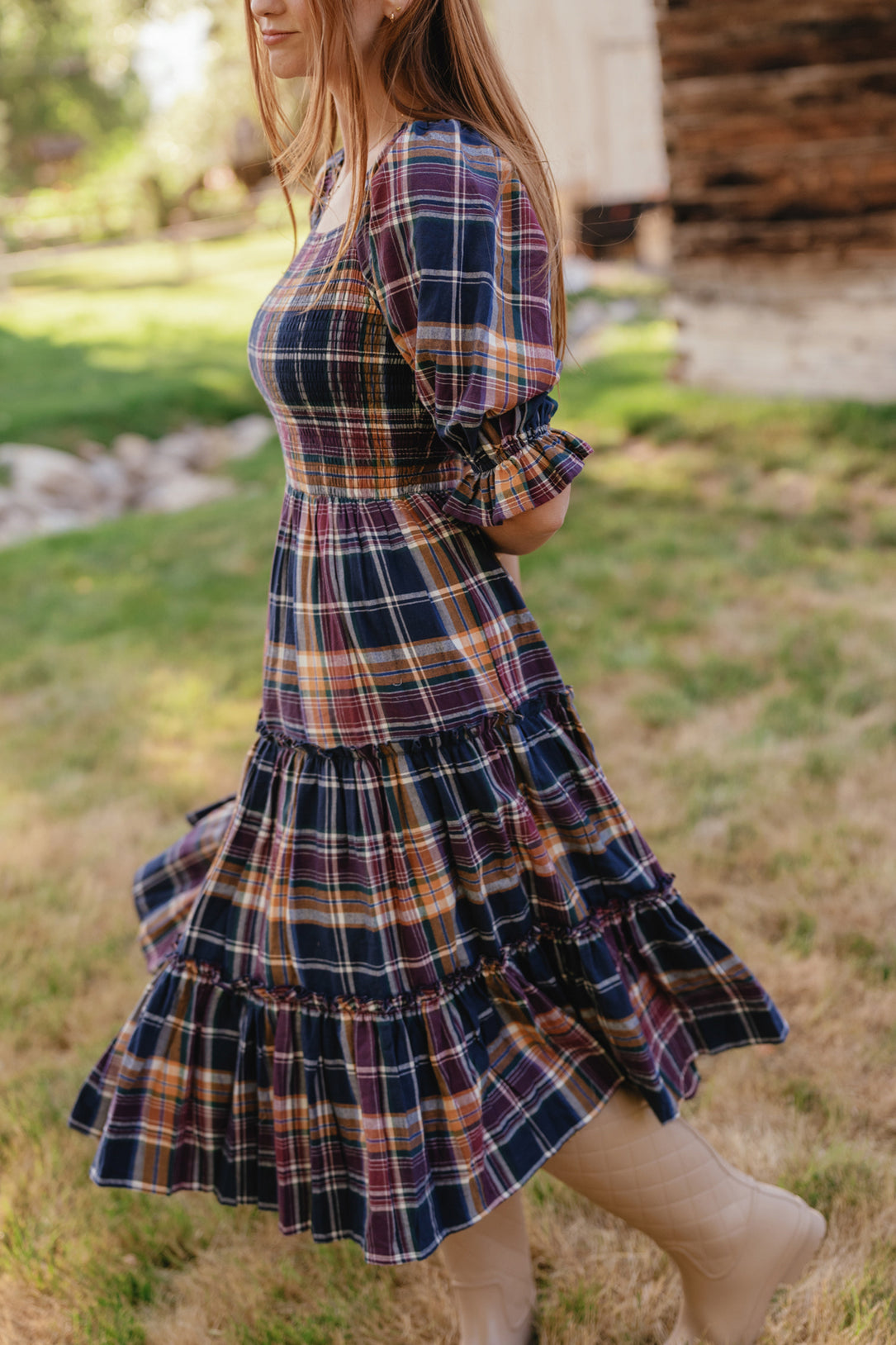 An expectant woman stands on grass near a rustic building, her long hair down and paired with light-colored knee-high boots. She wears the Ivy City Co Madeline Dress in Midnight Plaid—a maternity-friendly style crafted with tiers, a smocked bodice, puffy sleeves, and signature comfort and charm.