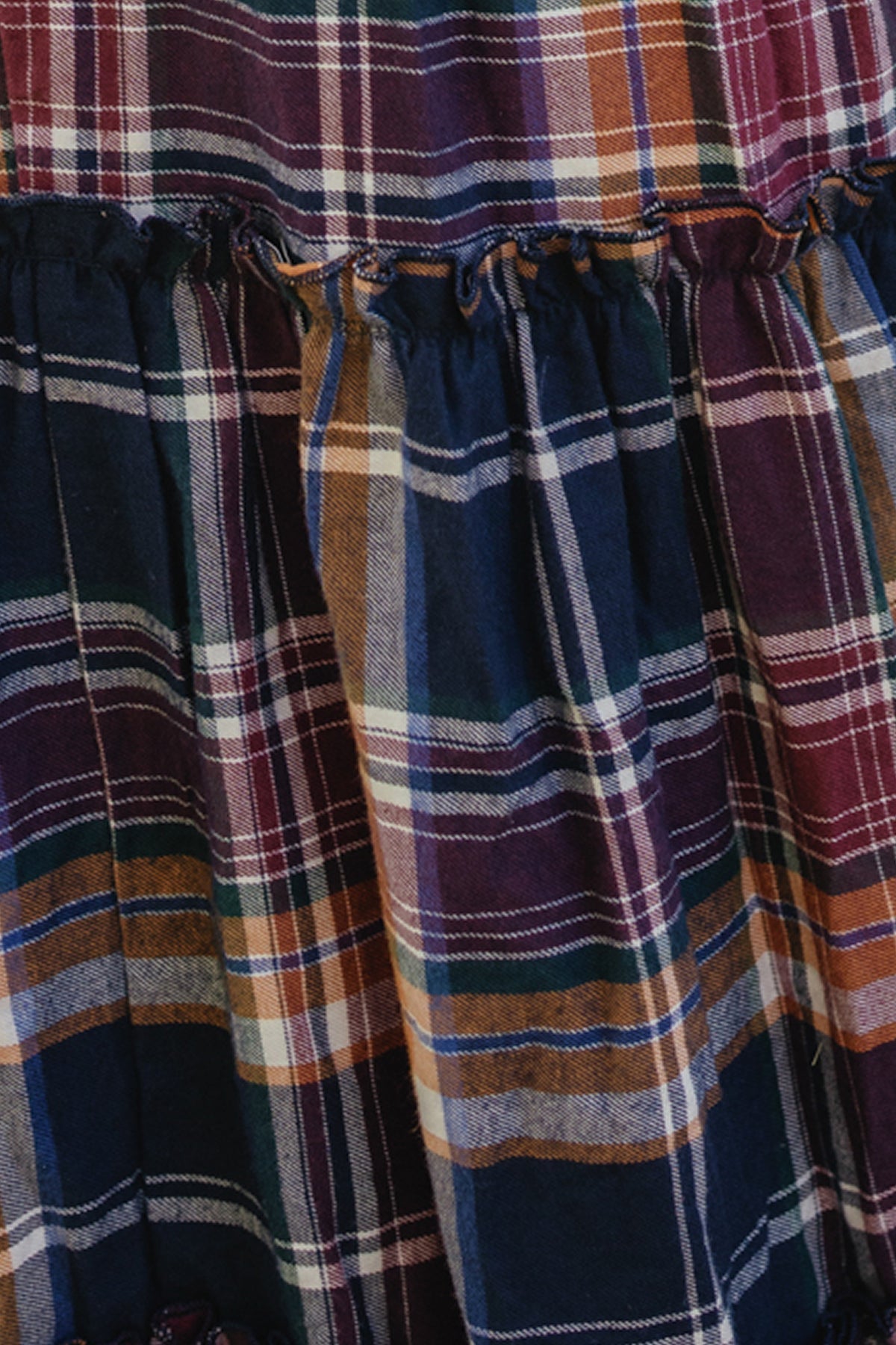 Close-up of fabric from the Ivy City Co Madeline Dress in Midnight Plaid, featuring a multicolored plaid pattern in shades of navy, orange, white, and purple, with gathered detailing along a seam and a smocked bodice for a comfortable, maternity-friendly fit.