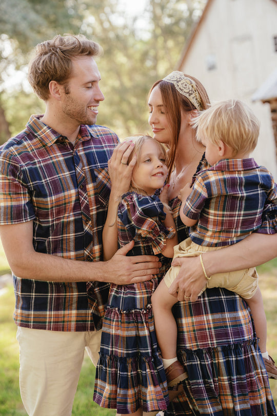 A family of four stands outdoors, smiling and embracing. They are all dressed in matching plaid outfits, with the mother elegantly wearing the Ivy City Co Madeline Dress in Midnight Plaid, featuring a smocked bodice and maternity-friendly design. The father holds his daughter while the mother holds her son, with trees and a building visible in the background.