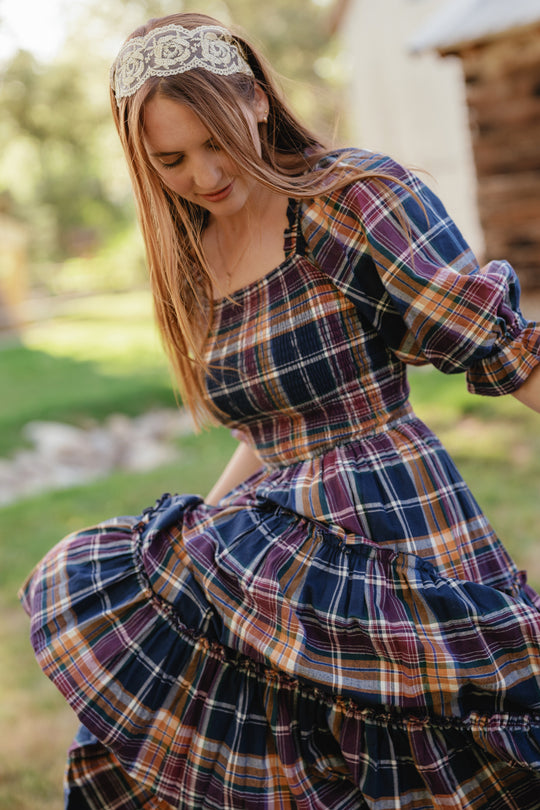 A woman with long hair, wearing a lace headband, twirls outdoors on the grass in the Madeline Dress in Midnight Plaid by Ivy City Co. Featuring a smocked bodice and puffed sleeves, this maternity-friendly dress is complemented by a backdrop of trees and a wooden building for a charming scene.