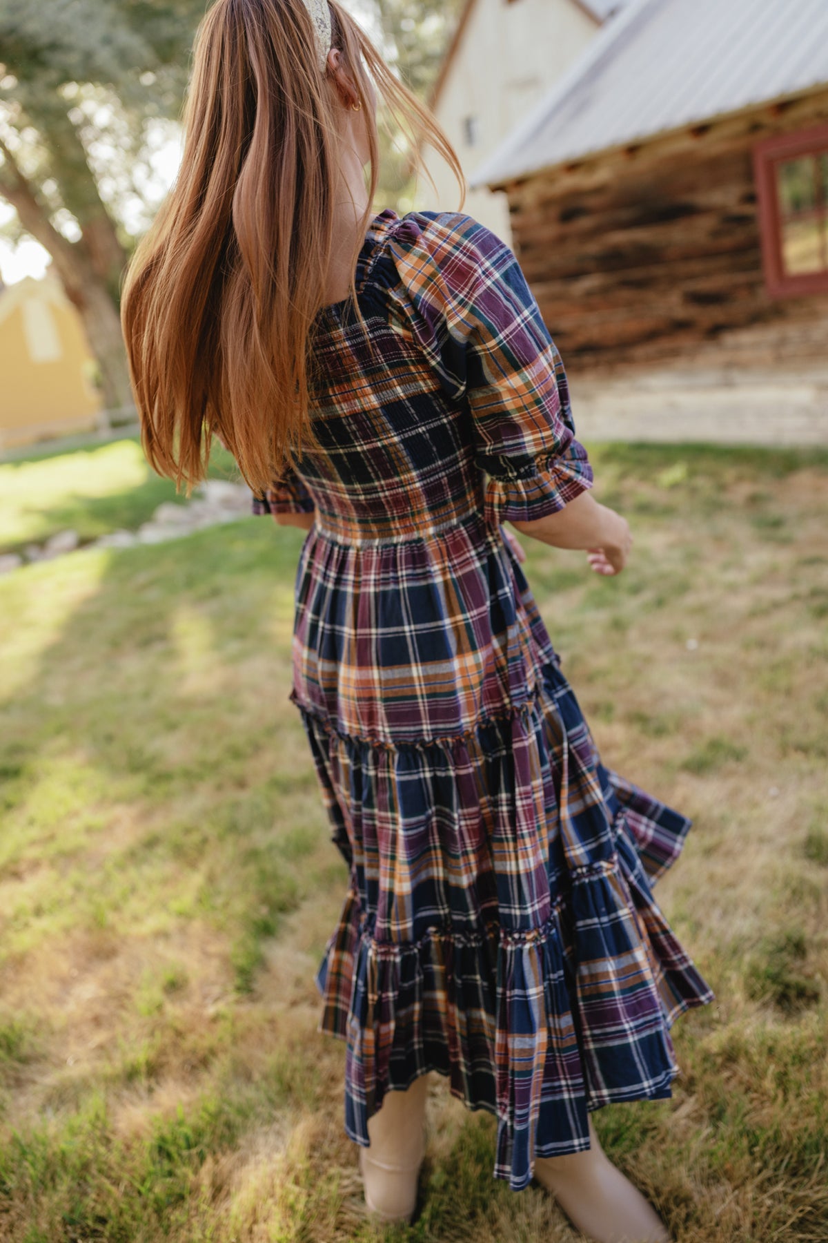 A girl with long hair, wearing the Ivy City Co Madeline Dress in Midnight Plaid—with its maternity-friendly smocked bodice—twirls outside on grass near rustic buildings, facing away from the camera.