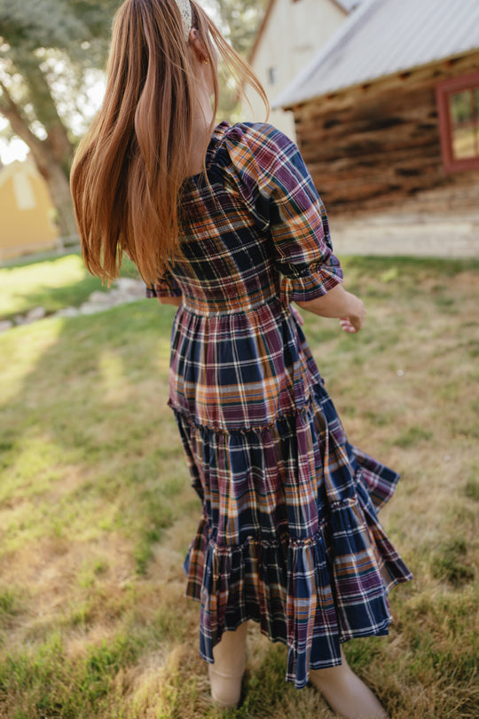 A girl with long hair, wearing the Ivy City Co Madeline Dress in Midnight Plaid—with its maternity-friendly smocked bodice—twirls outside on grass near rustic buildings, facing away from the camera.