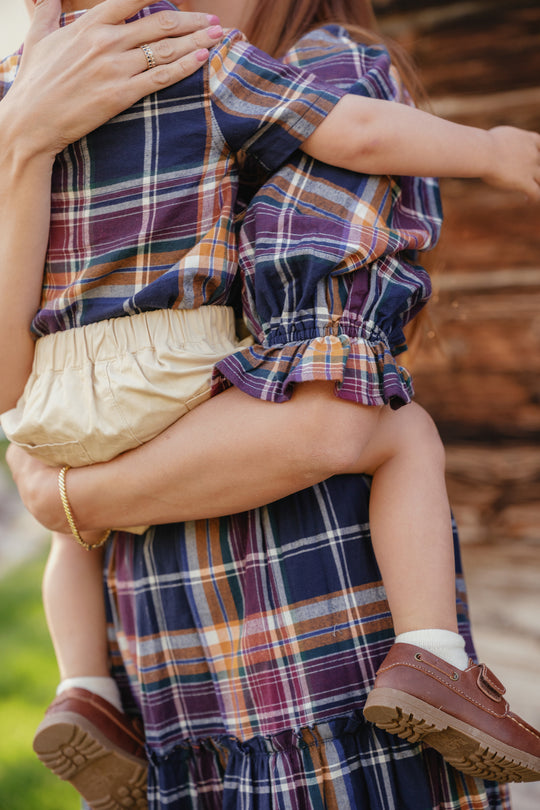 A woman wears the Ivy City Co Madeline Dress in Midnight Plaid, featuring a smocked bodice and a maternity-friendly design, as she holds a young child dressed in matching plaid. The child’s face is hidden, and they wear cream bloomers and brown shoes. They are outdoors by a wooden structure, their coordinated looks adding to the tranquil scene.