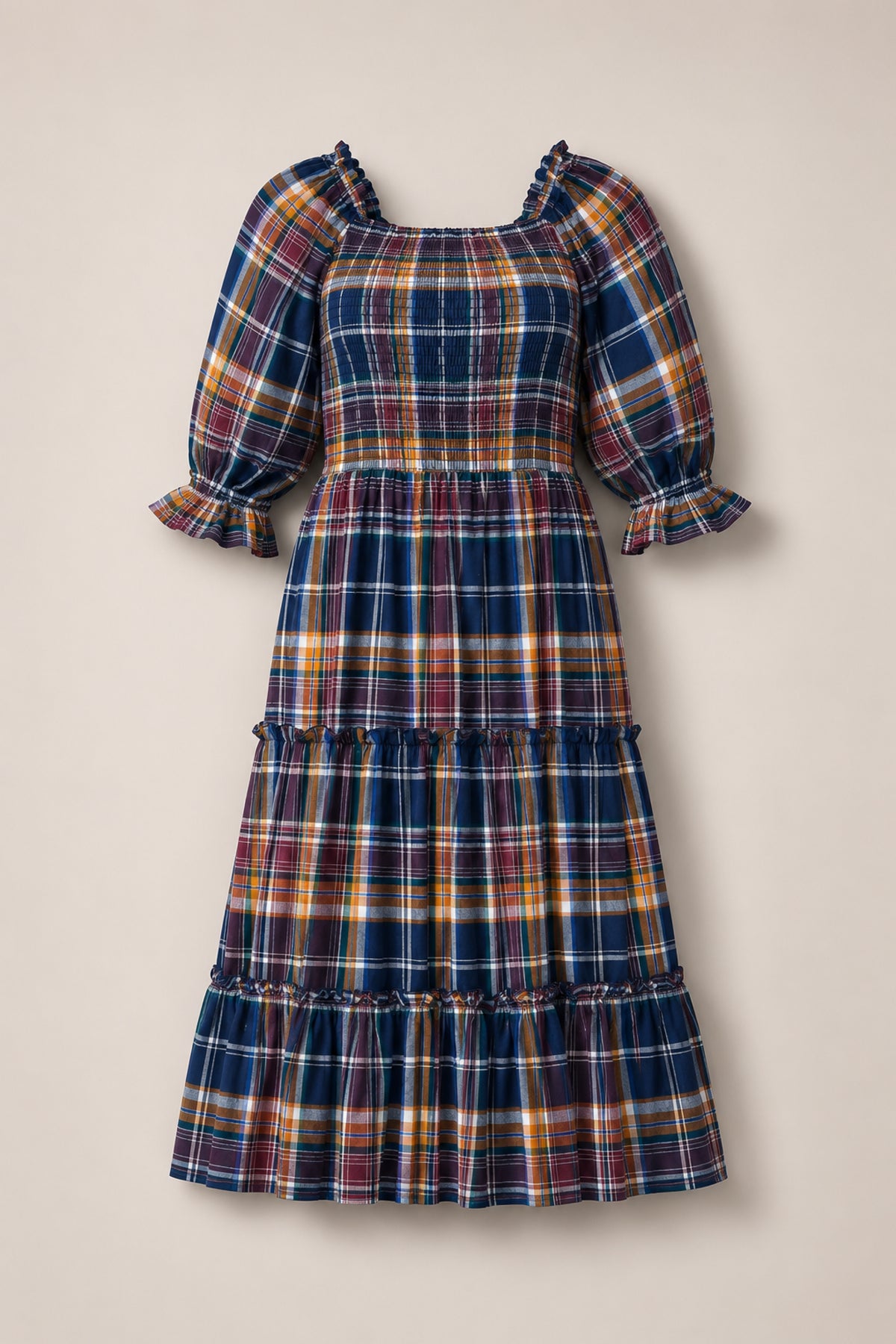 The Ivy City Co Madeline Dress in Midnight Plaid is a long, tiered dress with a smocked bodice for a comfortable, maternity-friendly fit. Featuring puffed sleeves with ruffled cuffs and a blue, yellow, red, and white plaid pattern, this dress is laid flat on a neutral background.