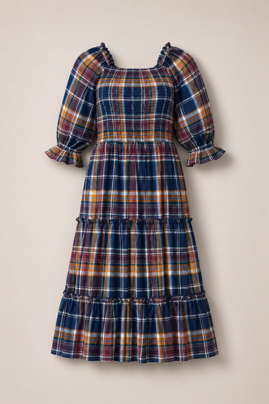 The Ivy City Co Madeline Dress in Midnight Plaid is a long, tiered dress with a smocked bodice for a comfortable, maternity-friendly fit. Featuring puffed sleeves with ruffled cuffs and a blue, yellow, red, and white plaid pattern, this dress is laid flat on a neutral background.