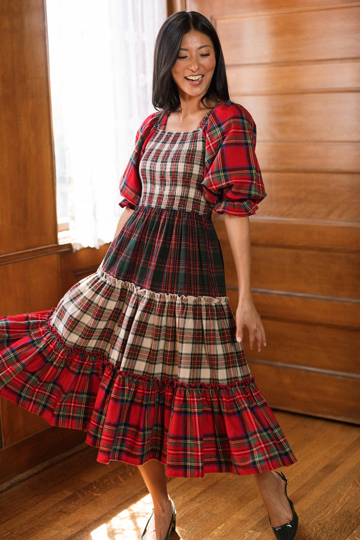 A woman smiles while twirling in the Madeline Dress in Mixed Plaid by Ivy City Co—a long dress with puffed sleeves and tiered layers, its vibrant red plaid perfect for Christmas family gatherings and designed to be maternity friendly. She stands in a sunlit, wood-paneled room, embodying festive elegance.
