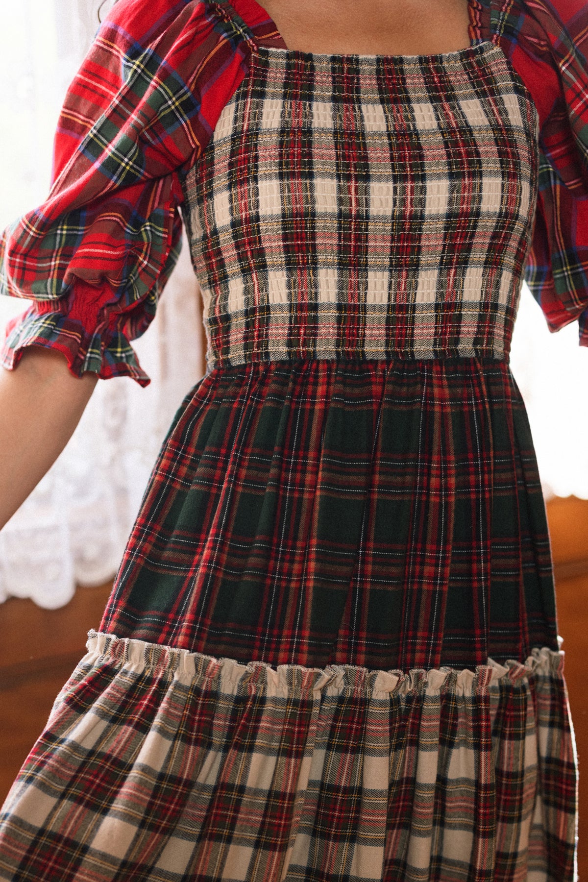 A person stands indoors near lace curtains, wearing the Ivy City Co Madeline Dress in Mixed Plaid. This maternity-friendly dress features red, green, and cream checkered patterns with puffed sleeves and a smocked bodice—making it perfect for cozy holiday gatherings.