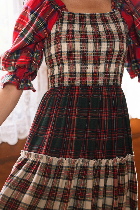 A person stands indoors near lace curtains, wearing the Ivy City Co Madeline Dress in Mixed Plaid. This maternity-friendly dress features red, green, and cream checkered patterns with puffed sleeves and a smocked bodice—making it perfect for cozy holiday gatherings.